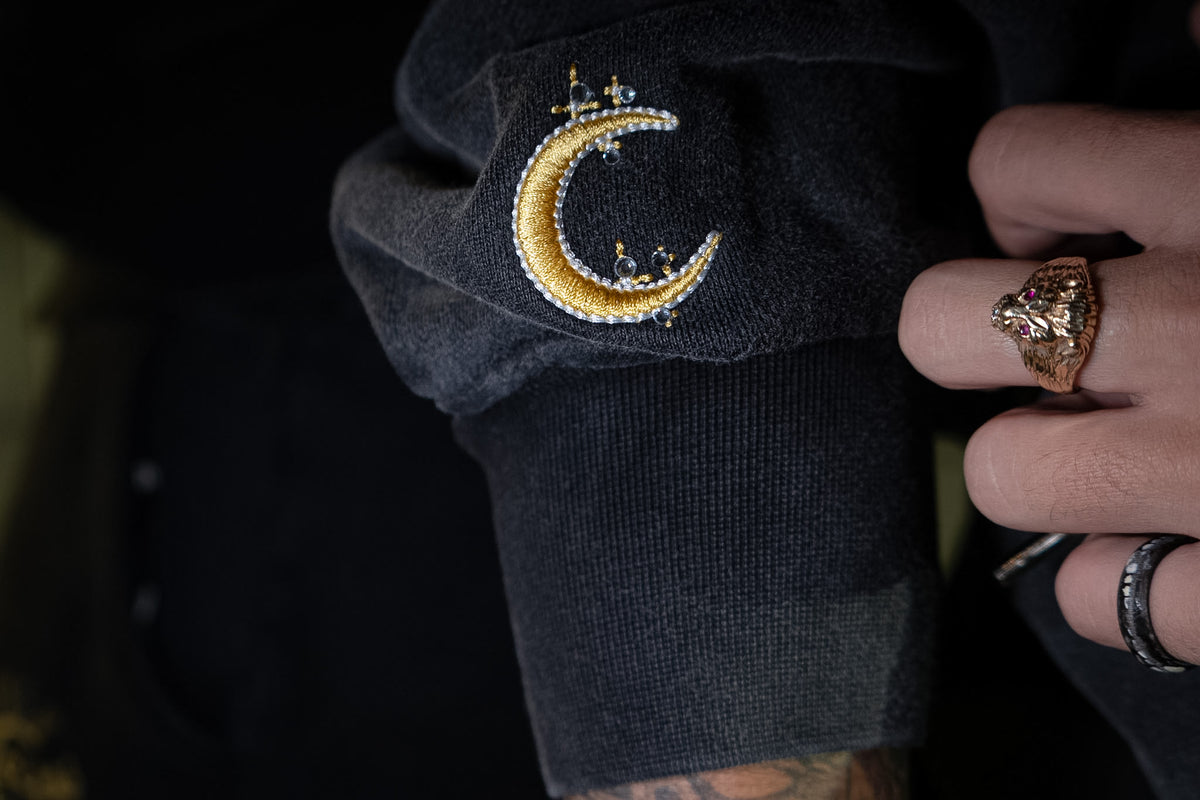 A hand at the cuff of the Dawn zip-up, gold crescent embroidery catching the light